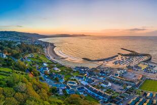 Blick von oben auf The Cobb und den Strand von Lyme Regis in Dorset bei Sonnenuntergang. Die goldenen Strahlen der untergehenden Sonne tauchen den malerischen Hafen und die Küste in warmes Licht.