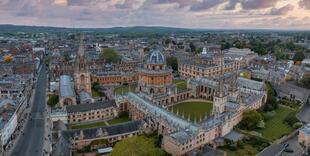 Luftbild der Stadt Oxford, mit ihren markanten historischen Gebäuden und den grünen Flächen. Die Universitätshochschulen und die berühmte Radcliffe Camera sind gut erkennbar, umgeben von den sanften Hügeln und dem Fluss.