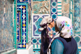  Zu sehen sind Frauen in traditioneller Kleidung vor der Nekropole Shah-i-Zinda in Samarkand, deren kunstvoll verzierte Mausoleen mit leuchtenden blauen Kacheln eine beeindruckende Kulisse bilden.