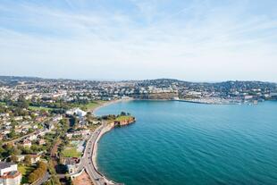 Blick von oben auf Torquay mit Meerblick. Die Küstenstadt erstreckt sich entlang der Bucht, mit weißen Gebäuden, üppigem Grün und einer glitzernden Wasseroberfläche.