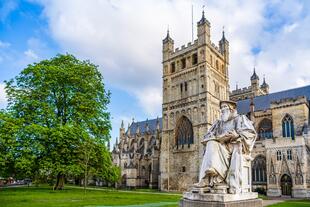 Die Statue von Richard Hooker in Exeter, mit dem historischen Gebäude im Hintergrund. Die Statue des Theologen steht auf einem Sockel, während das majestätische, gotische Gebäude der Kathedrale von Exeter im Hintergrund eine beeindruckende Kulisse bildet.