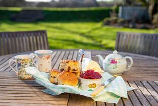 Ein Teller mit traditionellem Cream Tea, serviert auf einem Tisch. Der frische Scone ist großzügig mit clotted cream und Erdbeermarmelade belegt, begleitet von einer Tasse dampfendem Tee. 