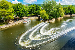 Blick auf die Pulteney Bridge über den Fluss Avon in Bath. Die beeindruckende Brücke mit ihren eleganten Bögen und historischen Gebäuden verläuft parallel zum Fluss, umgeben von grünen Ufern.