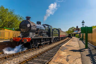 Es ist eine Dampflokomotive zu sehen. Diese ist berühmt für Sussex. Der historische Zug stößt weiße Dampfwolken aus.